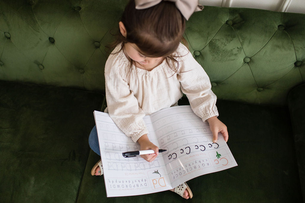 Child practicing writing letters in a dry erase alphabet book on a green couch, promoting interactive learning.